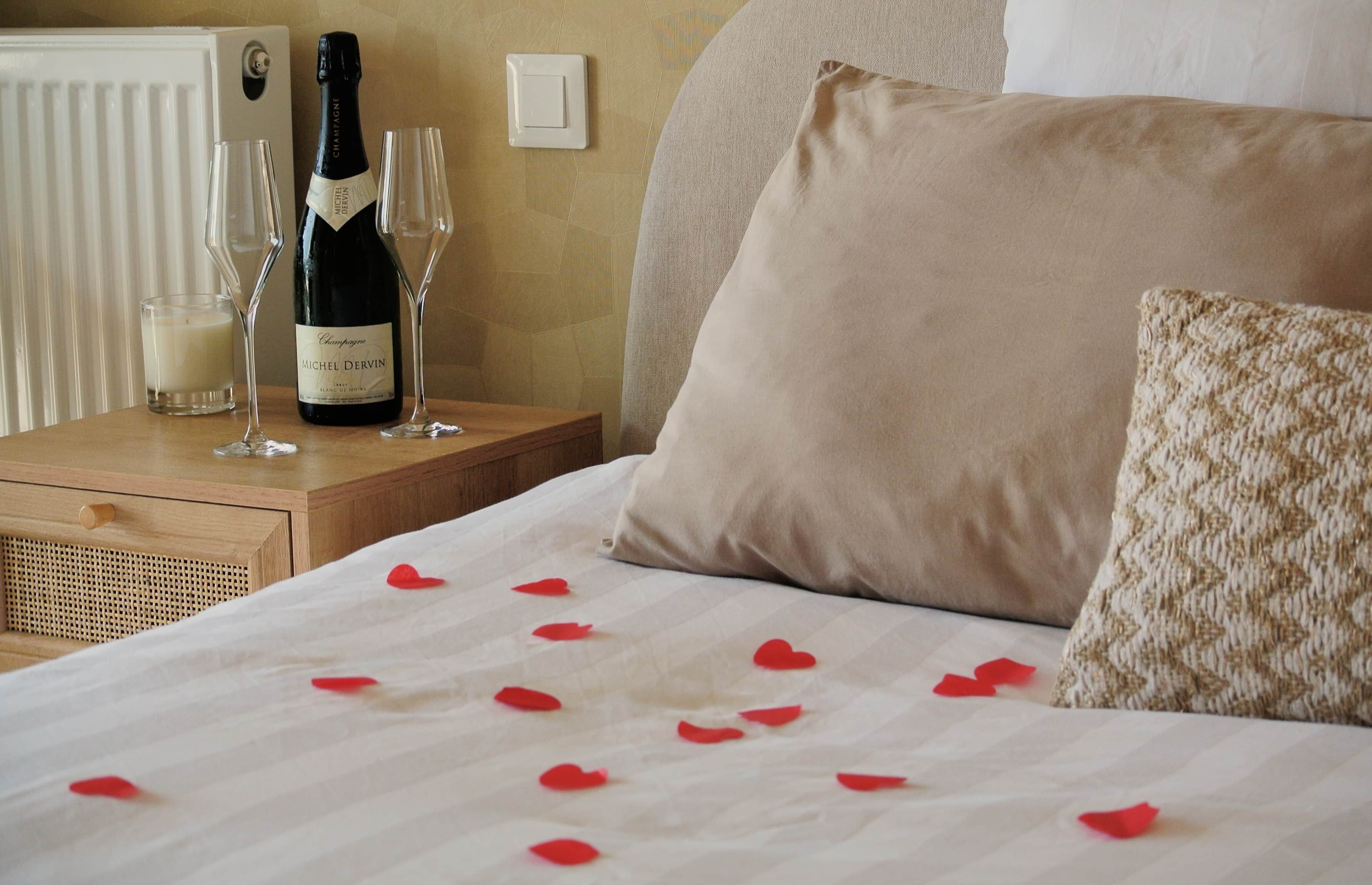Un lit soigneusement fait, parsemé de pétales de rose sur les draps, une bouteille de champagne et deux verres posés sur une table de chevet.