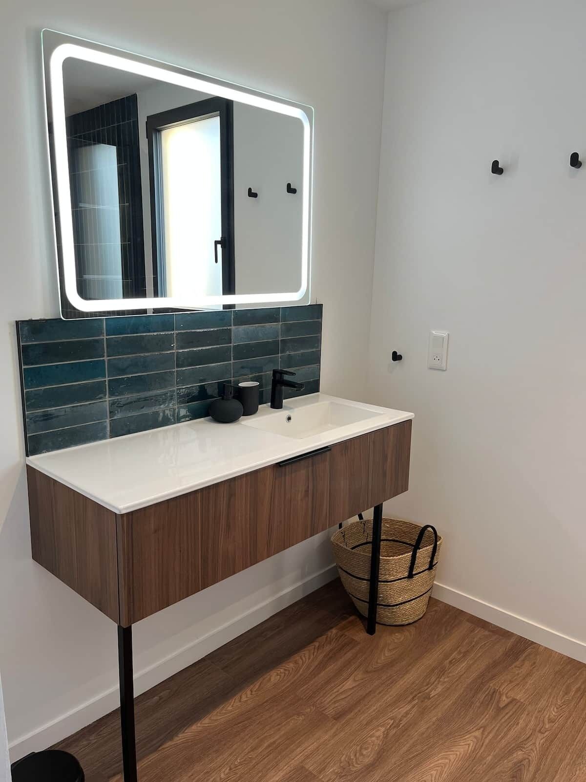 Un bagno con un lavandino bianco, un mobile bagno in legno scuro, uno specchio con cornice illuminata e uno schienale piastrellato blu.