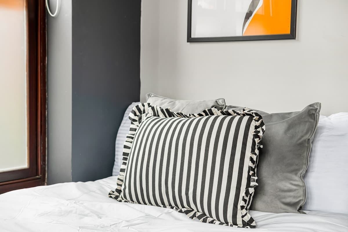 Una cama con ropa blanca, almohadas a rayas y un cojín gris.