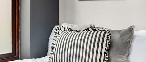 Una cama con ropa blanca, almohadas a rayas y un cojín gris.