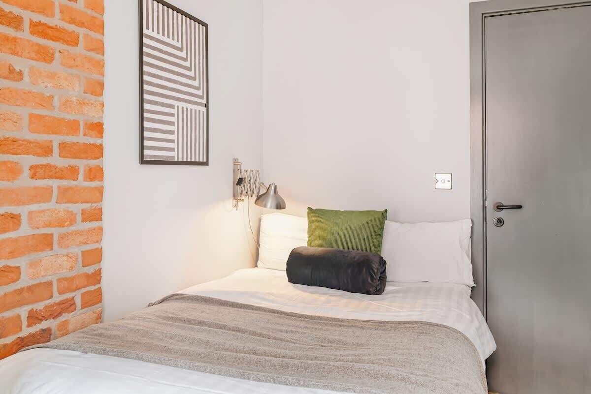A bedroom with a brick wall, a bed with white bedding, a green pillow, a black pillow, and a wall-mounted light fixture.