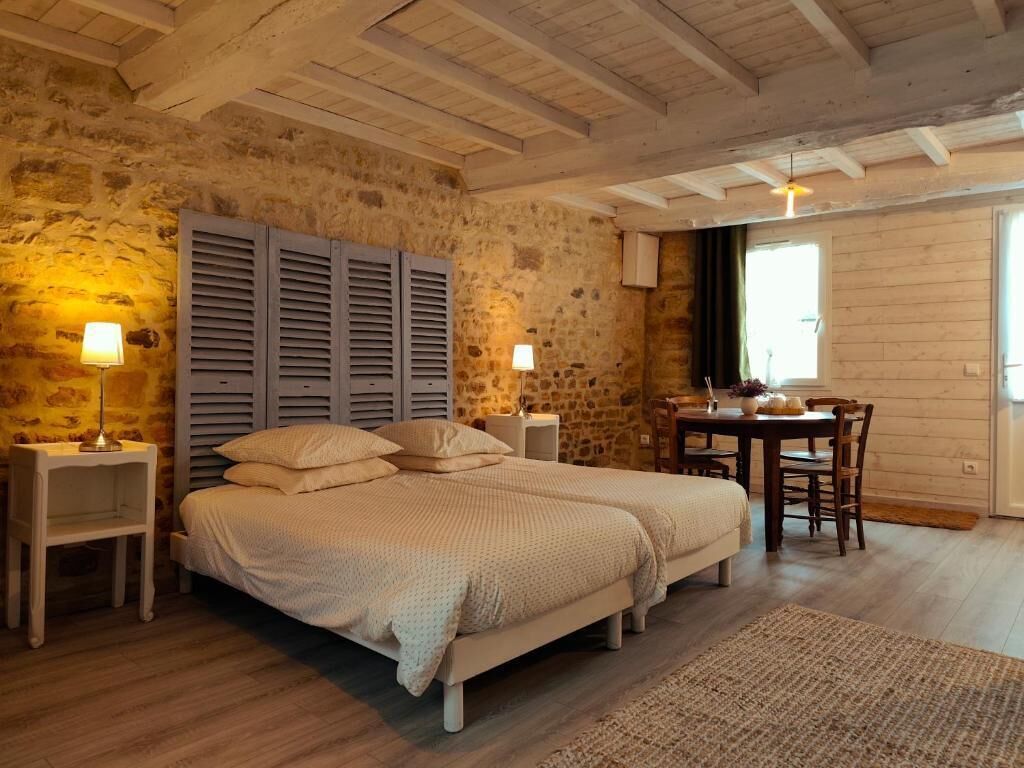 Ein Schlafzimmer mit zwei Betten, einer Steinwand, Holzbalken an der Decke, einem Esstisch mit Stühlen und einem Fenster.
