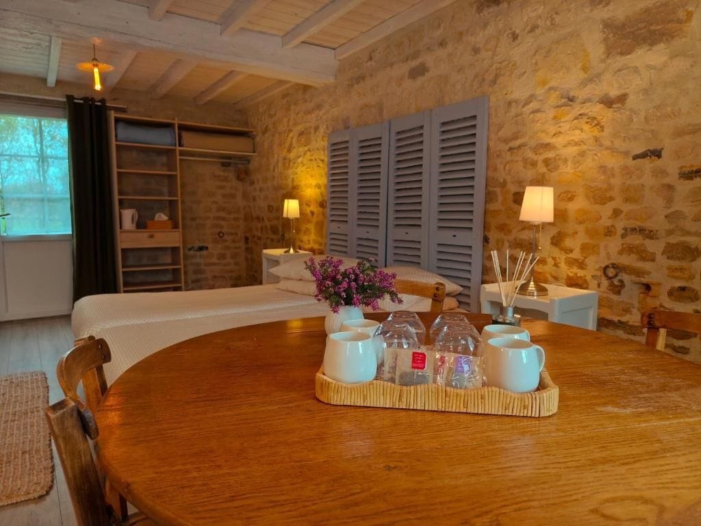 A room with a wooden dining table set with white cups and a tray, a bed with white linens, a stone wall, and a window with curtains.