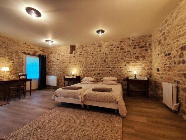 A bedroom with a stone wall, a double bed, bedside tables with lamps, a desk with a chair, and a radiator.