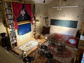 A cozy room with a large flat-screen TV, bookshelves, a wooden desk, a patterned rug, and multiple chairs.