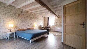 Een slaapkamer met een stenen muur, houten balken, een bed, een tafel met stoelen en een houten deur.