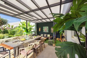 A modern outdoor dining area with a long table, chairs, and a bar. There are large green plants and a view of the surrounding garden. - Dream Vacation Home with Ocean View, Private Pool, and Garden in Kournas (Kournas Natures Nest)