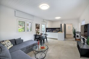 A modern living room with a grey sofa, a glass coffee table with fruit, and a dining area with black chairs. - Dream Vacation Home with Ocean View, Private Pool, and Garden in Kournas (Kournas Natures Nest)