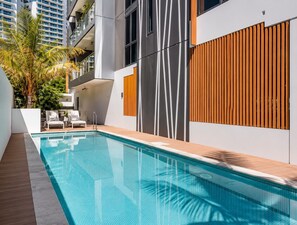 Piscina exterior moderna con superficie de agua azul cristalina, rodeada por un muelle de madera y un muro blanco con listones de madera verticales.