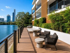 Un espacio al aire libre frente al agua, con mesas y sillas, con vistas a un cuerpo de agua y el horizonte de la ciudad.