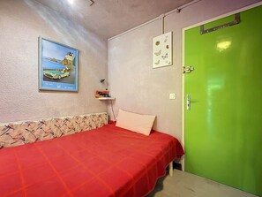 Ein Zimmer mit einem Bett, einer grünen Tür, einem gerahmten Bild an der Wand und einer kleinen Ablagefläche mit Gegenständen.
