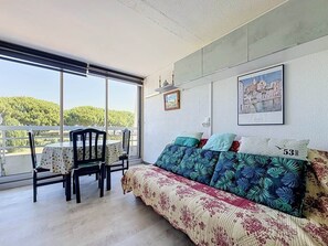 Ein Zimmer mit Balkon, ein Sofa mit gemusterten Kissen, ein Tisch mit Stühlen und ein gerahmtes Bild an der Wand.