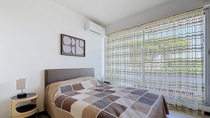 Ein Schlafzimmer mit Bett, Nachttisch, Klimaanlage und einem Fenster mit Vorhang.
