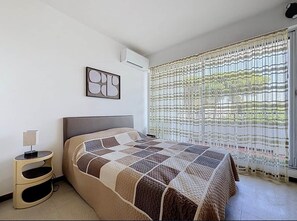 A bedroom with a bed, bedside table, air conditioning unit, and a window with a curtain. - A Pleasant Marina in the DRC (Le Grau-du-Roi)