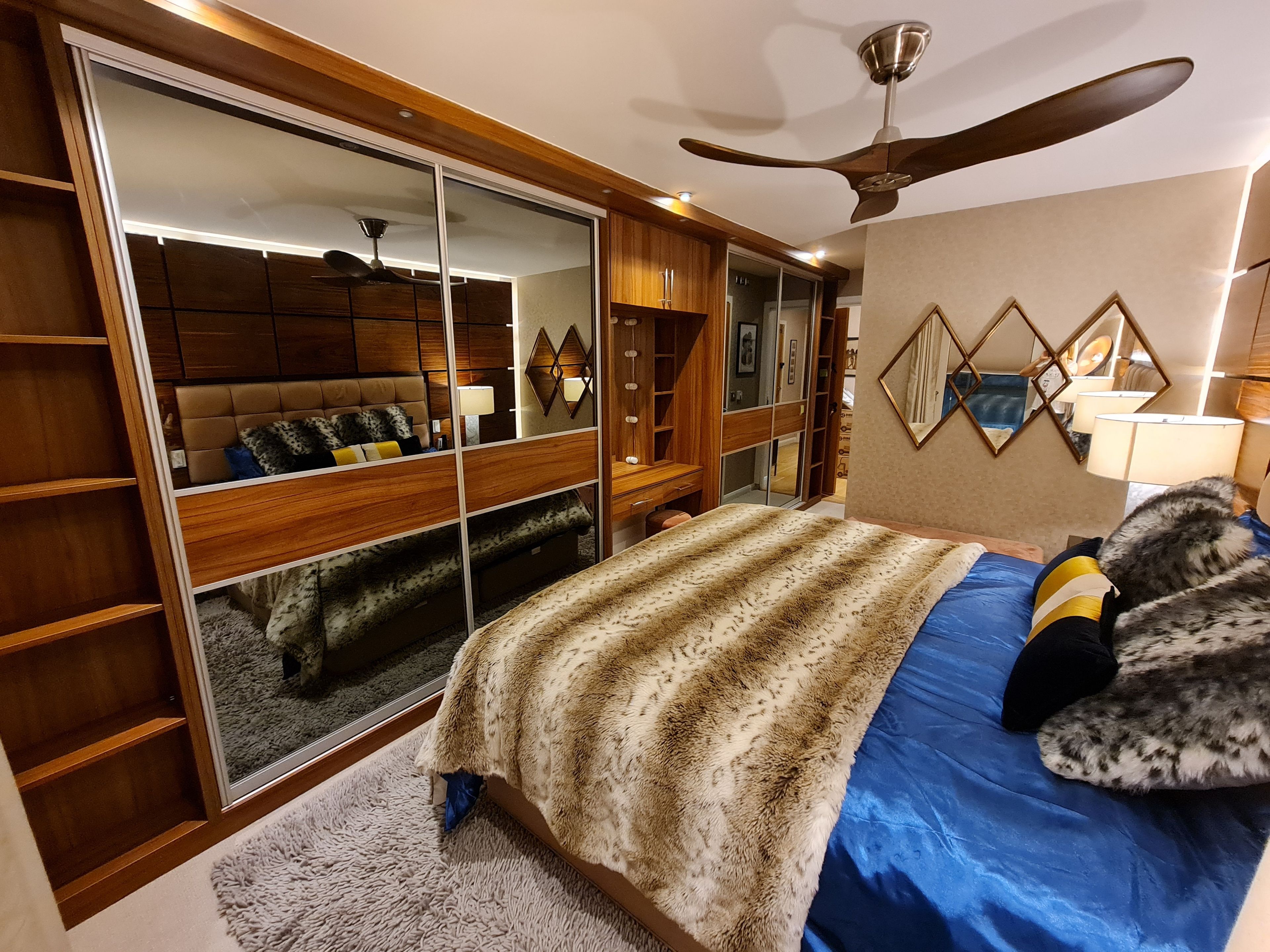 A bedroom with a large bed, a ceiling fan, a wardrobe, and a mirror.
