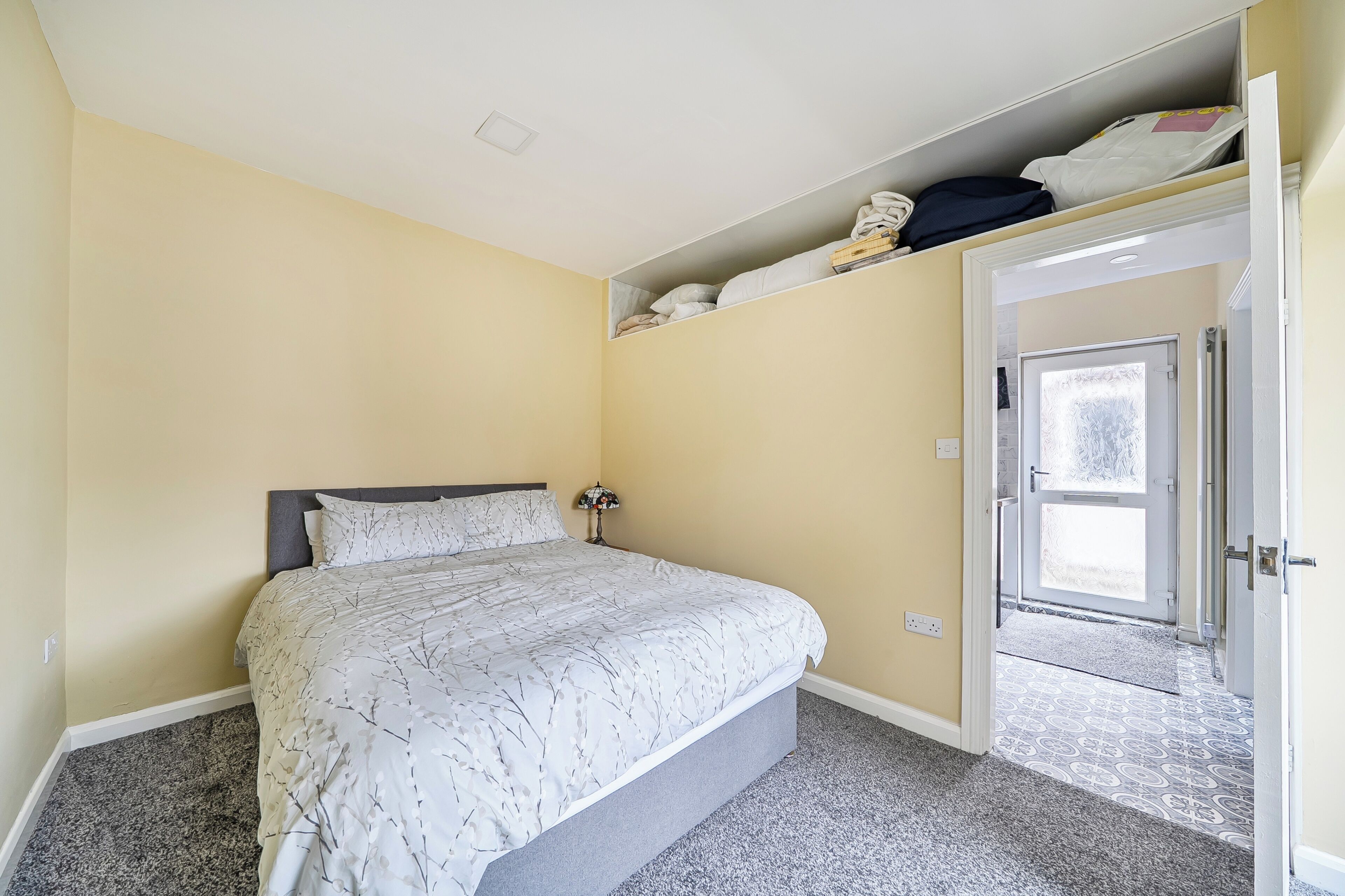 A bedroom with a bed, a nightstand, a lamp, and a door leading to another room.
