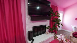 Un salón decorado en tonos rosas, con un televisor de pantalla plana sobre la chimenea, una planta en maceta y un jarrón decorativo.