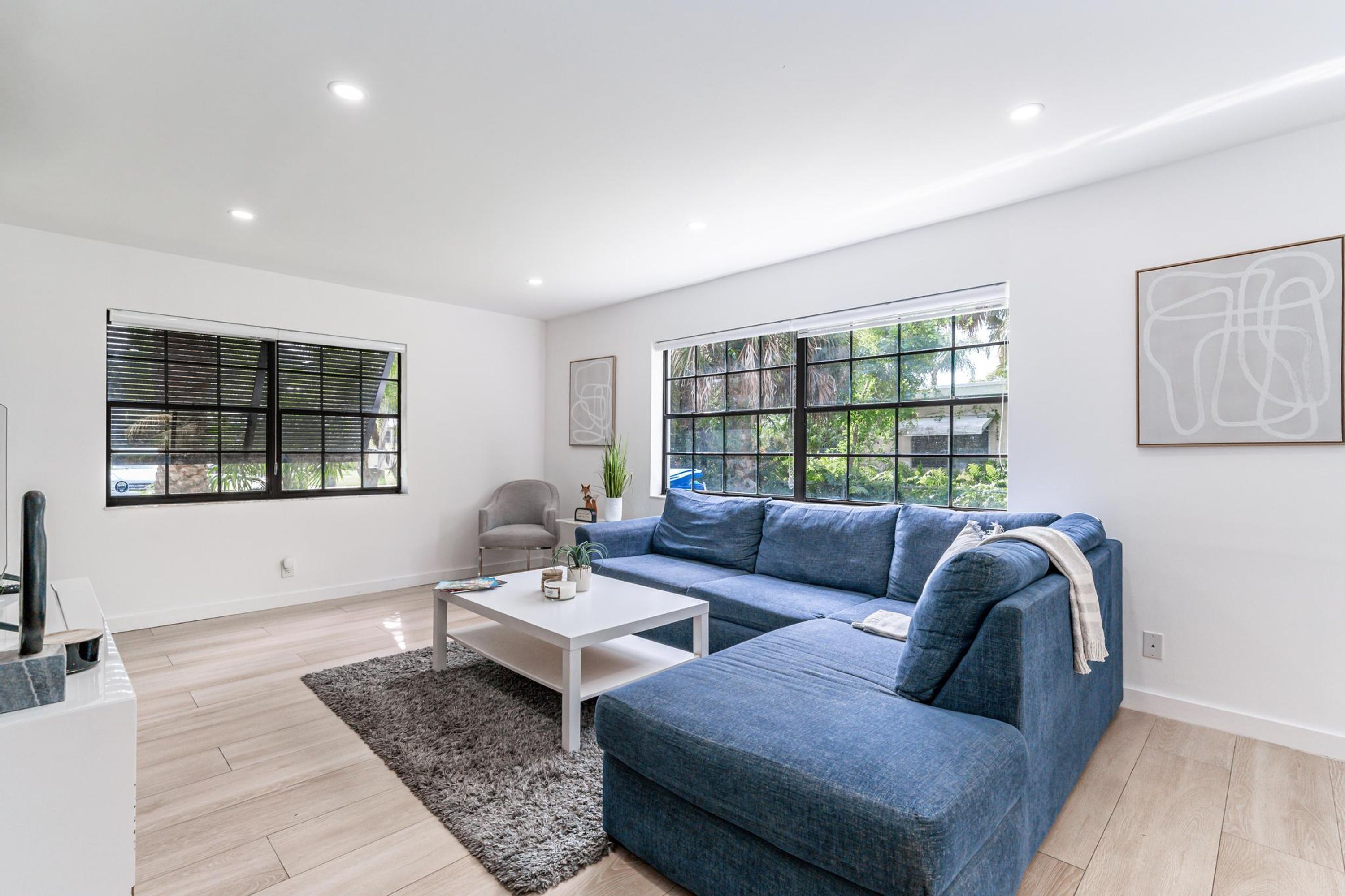 Una sala de estar moderna con un sofá azul, una mesita de centro blanca y dos ventanas con persianas.