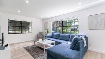Una sala de estar moderna con un sofá azul, una mesita de centro blanca y dos ventanas con persianas.