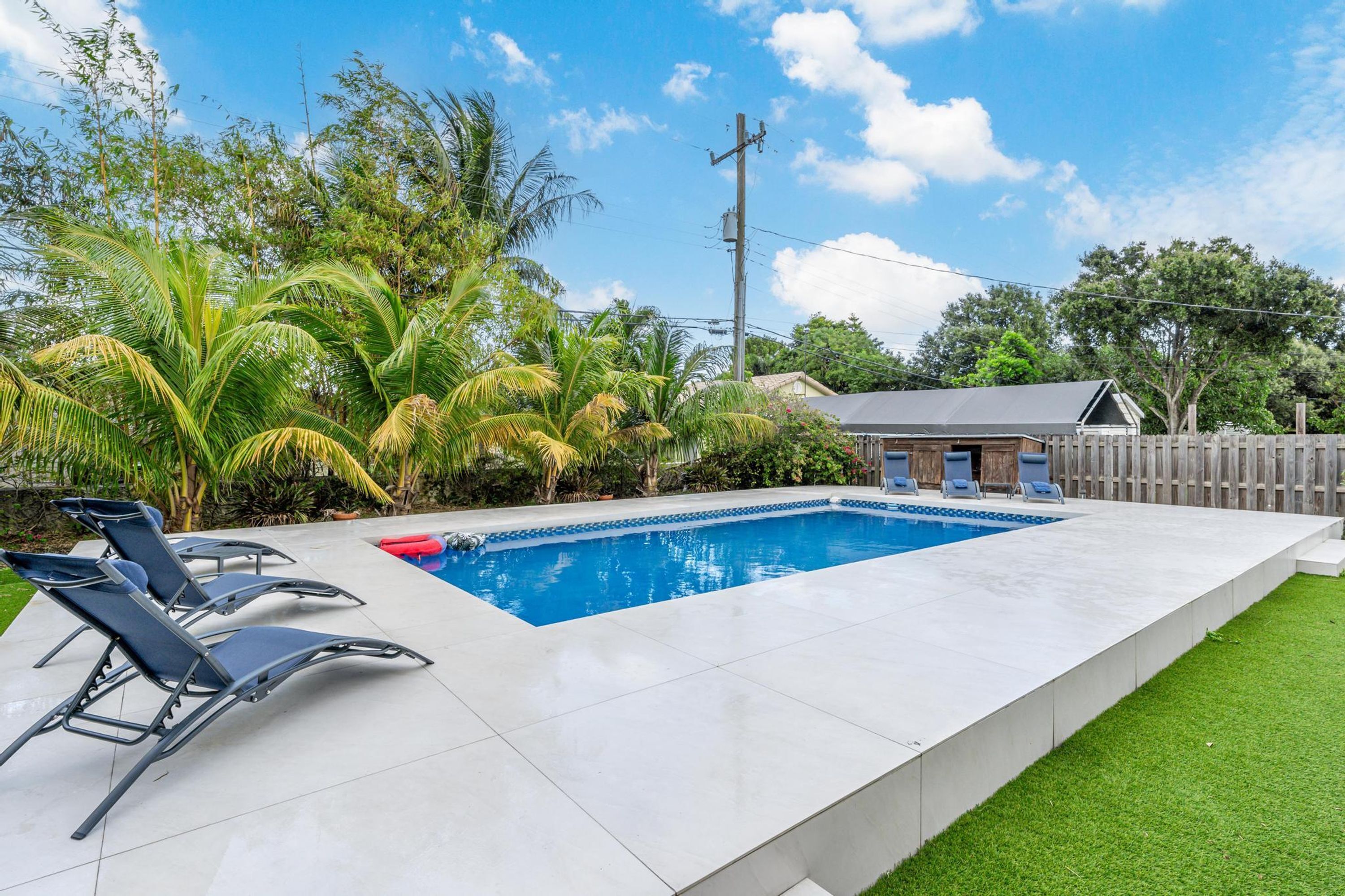 Una piscina rectangular con un borde de baldosas, rodeada de palmeras y una valla de madera.