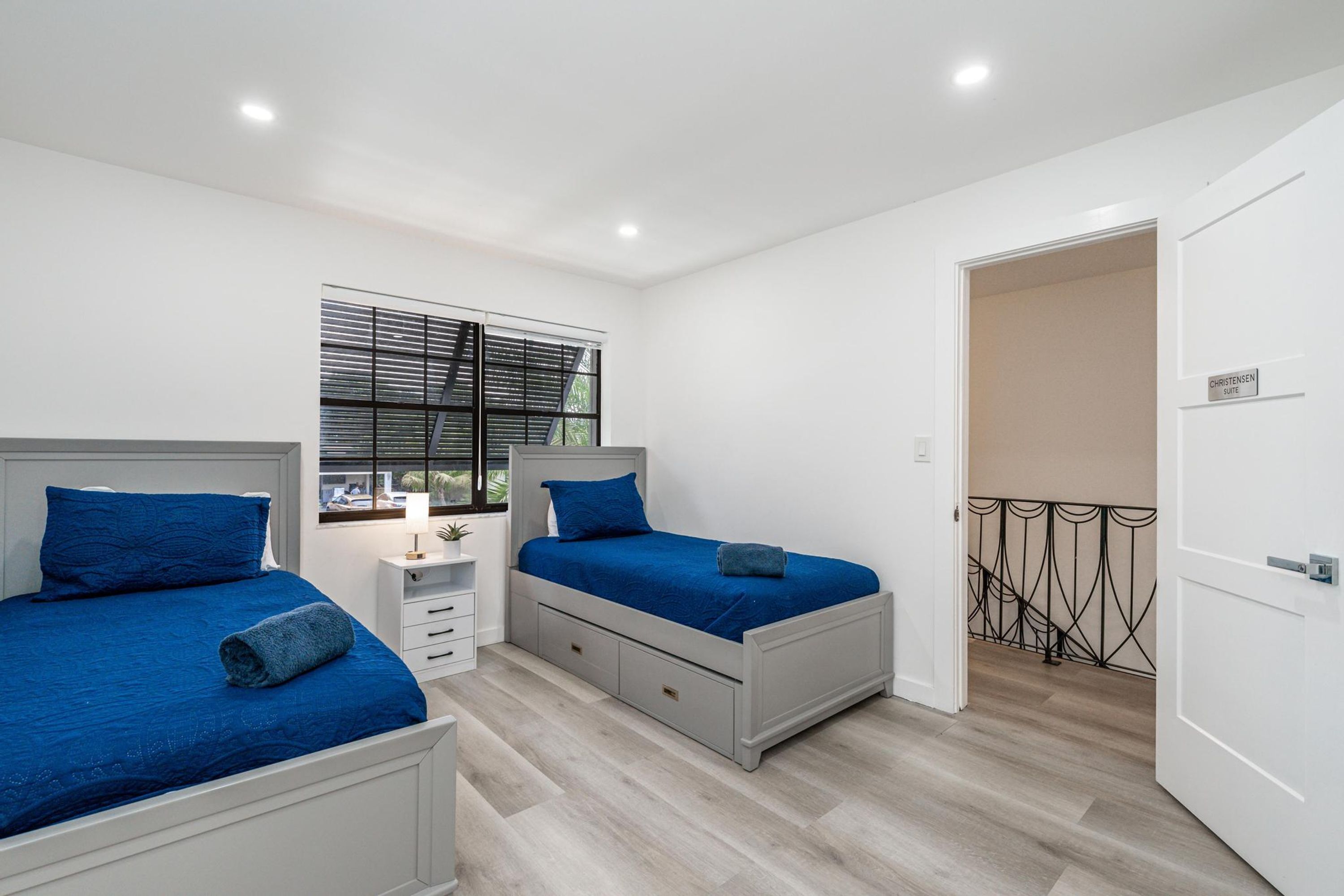 Un dormitorio con dos camas, una ventana con persianas, una mesita de noche y una puerta a un balcón.