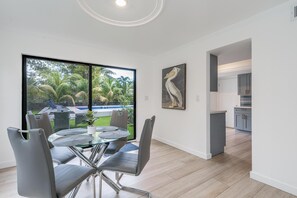 A modern kitchen with grey cabinets, a dining area with a glass table and grey chairs, and a large window with a view of a pool and greenery. - Fox Palace Pool House (Fort Lauderdale)