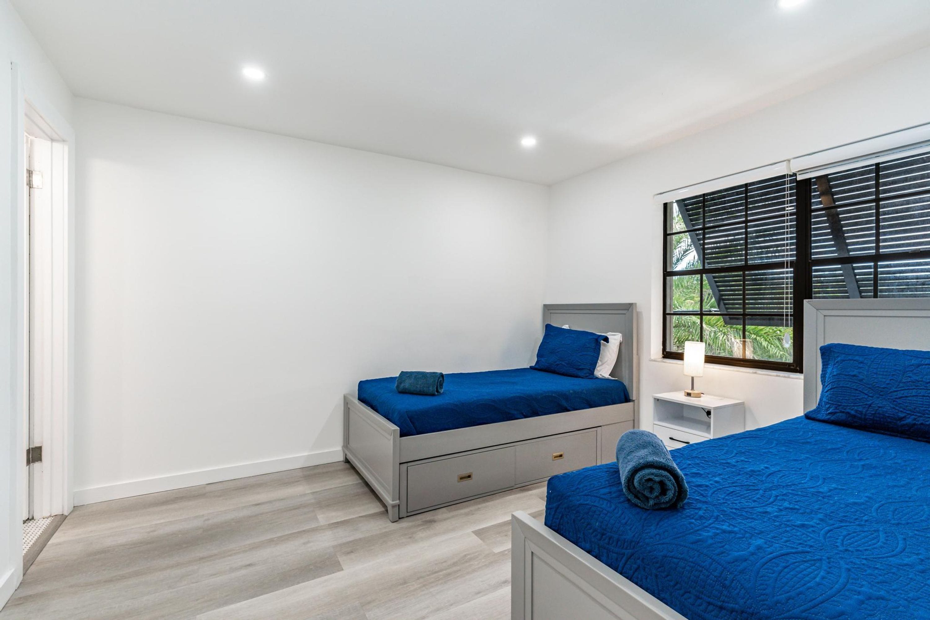 Un dormitorio con dos camas, una ventana con persianas, una mesita de noche con una lámpara y una puerta.