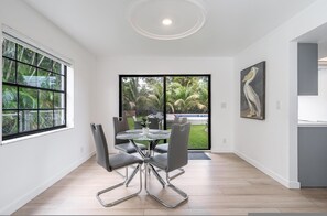 Un comedor moderno con una mesa de vidrio, sillas metálicas y vista a un jardín a través de puertas de cristal.
