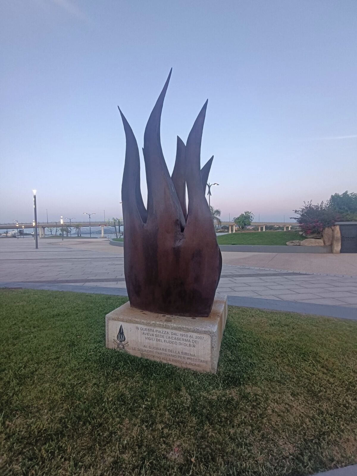 En skulptur i metall på ett offentligt torg, med en plakett, en bro i bakgrunden och en klar himmel.