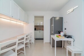 A modern kitchen with white cabinets, a stainless steel refrigerator, and a dining area with a white table and chairs. - Domo Archea (Olbia)