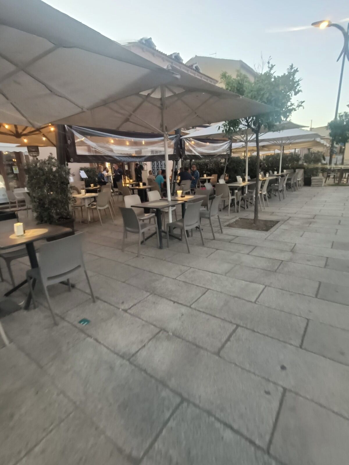 En udendørs café med borde, stole og parasoller, samt en belagt sti.