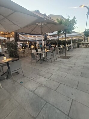 An outdoor café with tables and chairs, covered by umbrellas, and a paved walkway. - Domo Archea (Olbia)
