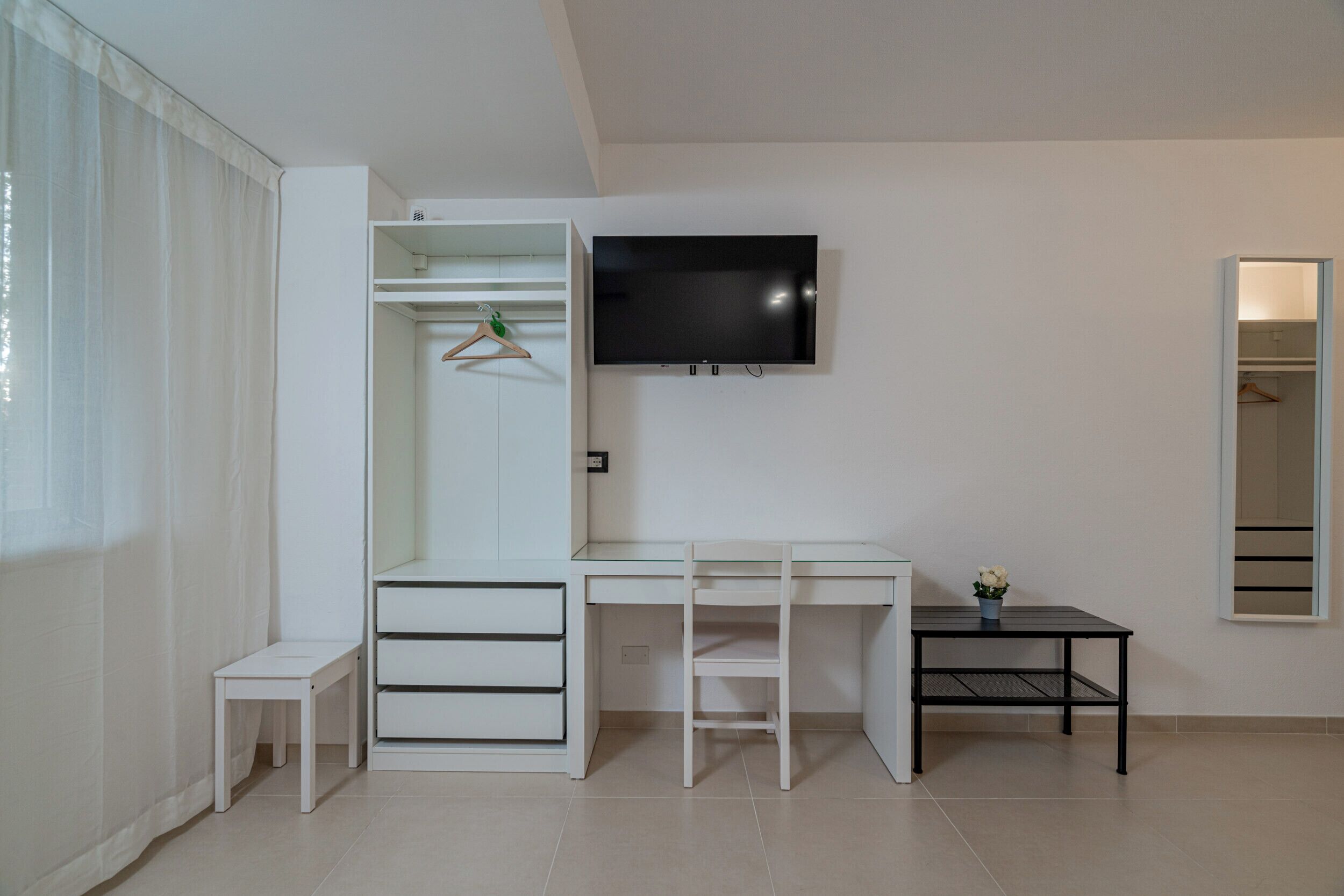 Ein moderner, minimalistischer Raum mit weißem Schreibtisch, einem an der Wand montierten Fernseher, einem kleinen Tisch mit Topfpflanze und einem Kleiderschrank mit Kleiderstange.