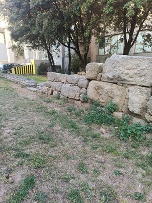 Un mur de pierre avec une zone herbeuse devant, une voiture garée et un bâtiment avec des fenêtres en arrière-plan.