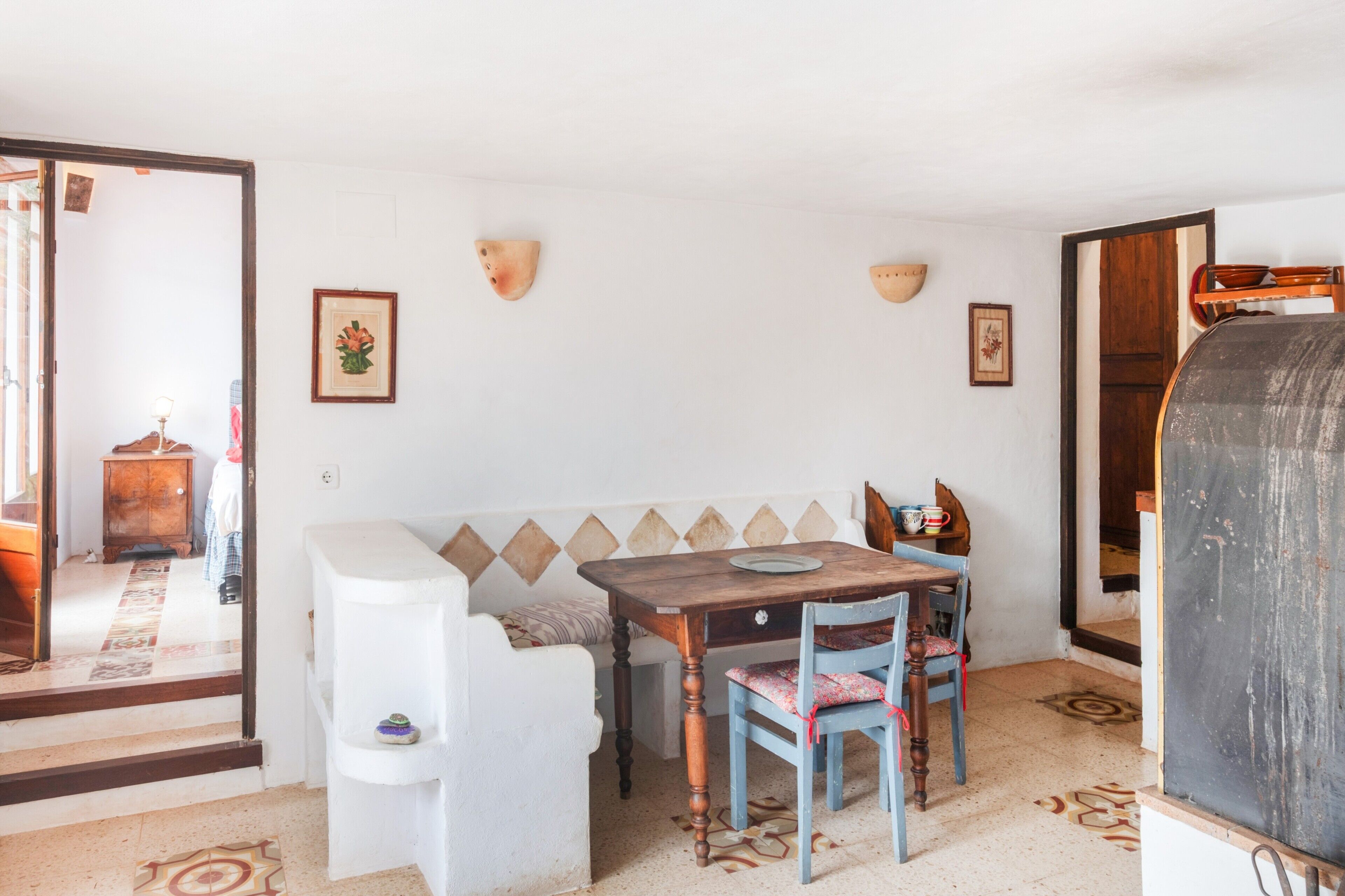 A room with a white sofa, a wooden table, two chairs, a wall-mounted shelf, and a door leading to another room.