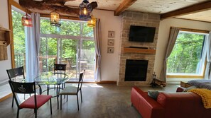 A cozy living room with a glass dining table, red and black chairs, a red sofa, a fireplace, and large windows with a view of trees. - Spa & Sauna | Plume Boreale (Saint-Alexis-des-Monts)