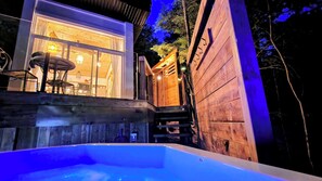 A wooden cabin with a hot tub, a dining area with a table and chairs, and a staircase leading to a deck. - Spa & Sauna | Plume Boreale (Saint-Alexis-des-Monts)