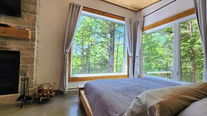 A bedroom with a large window, a fireplace, and a view of trees. - Spa & Sauna | Plume Boreale (Saint-Alexis-des-Monts)