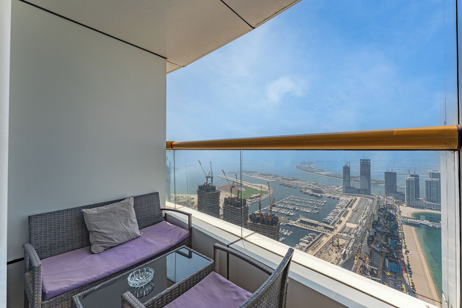 Ein Balkon mit einer Rattan-Couch und einem Glastisch, mit Blick auf eine Stadtlandschaft mit Gebäuden und einem Yachthafen.