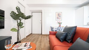 A modern living room with a sofa, a coffee table with magazines, a plant, and a dining area with a table and chairs.