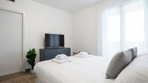 Habitación de hotel con cama, televisor sobre un soporte, planta en maceta y ventana con cortinas.