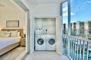 A bedroom with a bed, nightstand, and a view of a cityscape through a balcony. - Casa Marina 644 - Bay View (Fort Myers Beach)