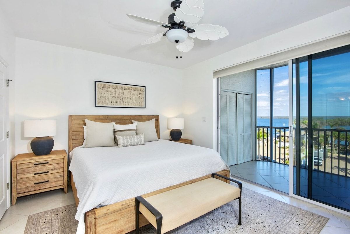 A bedroom with a bed, bedside tables, a ceiling fan, a large window, and a balcony with a view.