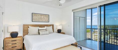 A bedroom with a bed, bedside tables, a ceiling fan, a large window, and a balcony with a view.