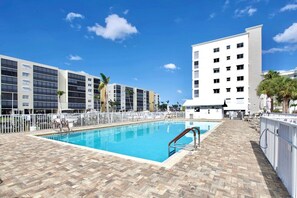 A swimming pool with a tiled deck, surrounded by a white fence, and adjacent to a multi-story building with balconies. - Casa Marina 644 - Bay View (Fort Myers Beach)