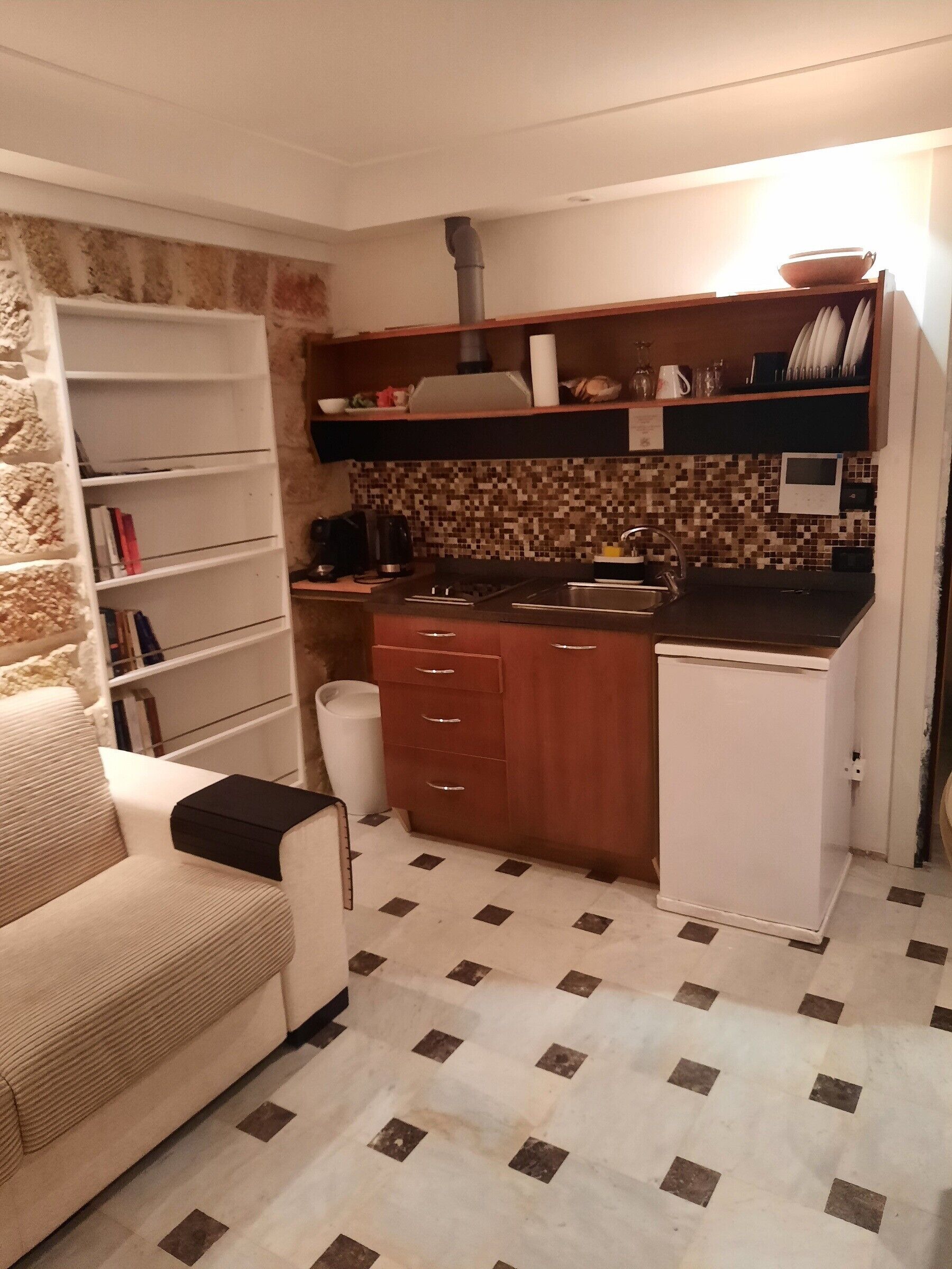 A compact kitchen with a microwave, sink, and a small refrigerator. There is a bookshelf with books and a white trash bin. The floor has a patterned tile design.