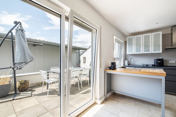 A modern kitchen with a large window, a dining area with white chairs, and a small outdoor seating space.