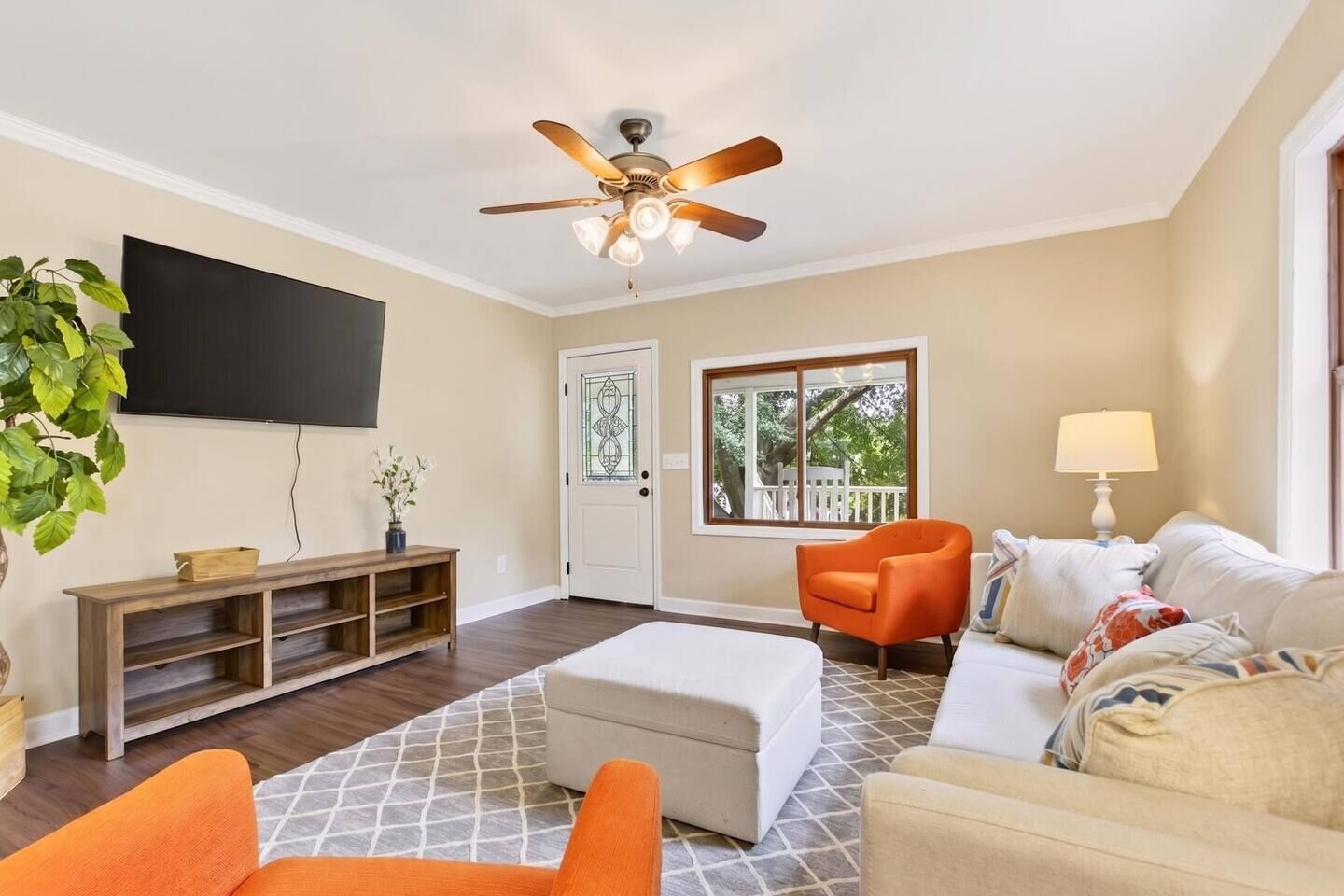 A living room with a ceiling fan, a TV, a wooden entertainment unit, a white sofa, an orange armchair, and a window with a view of trees.
