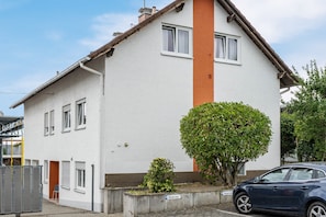 Un edificio residencial de dos pisos con fachada blanca, un muro de acento marrón y un árbol pequeño en la entrada.