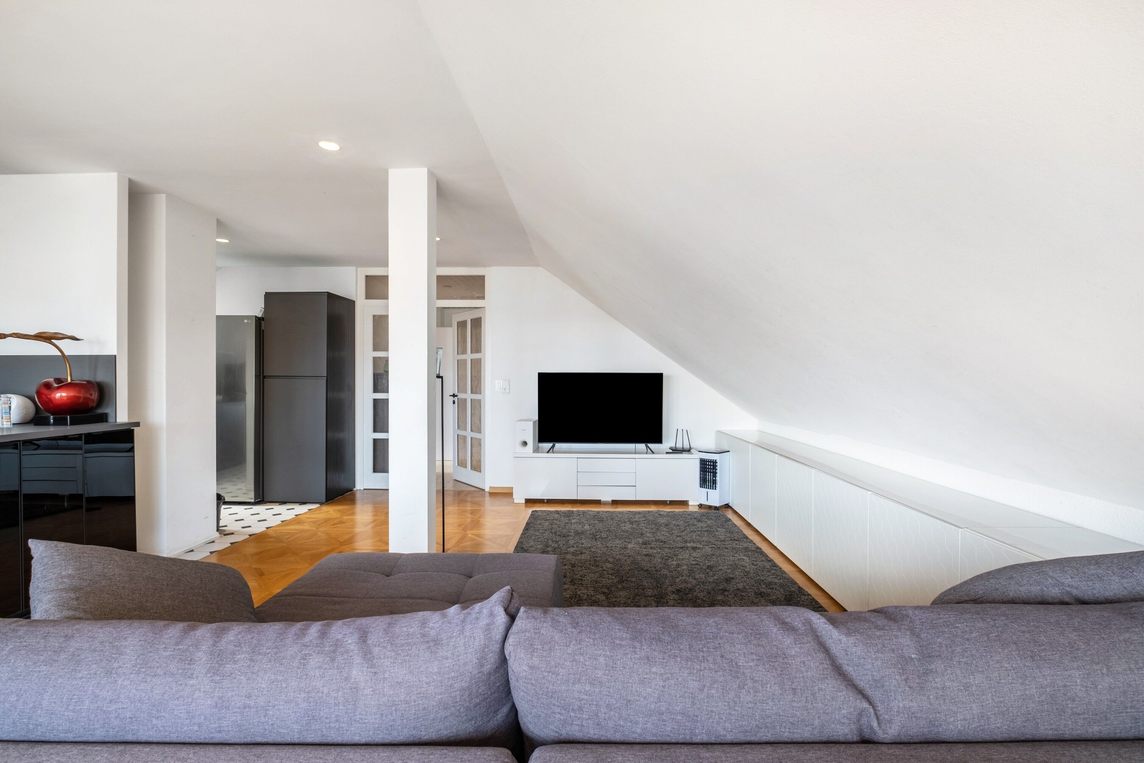 A modern living room with a high ceiling, a flat-screen TV mounted on a white cabinet, a grey sofa, and a dining area with a kitchen in the background.